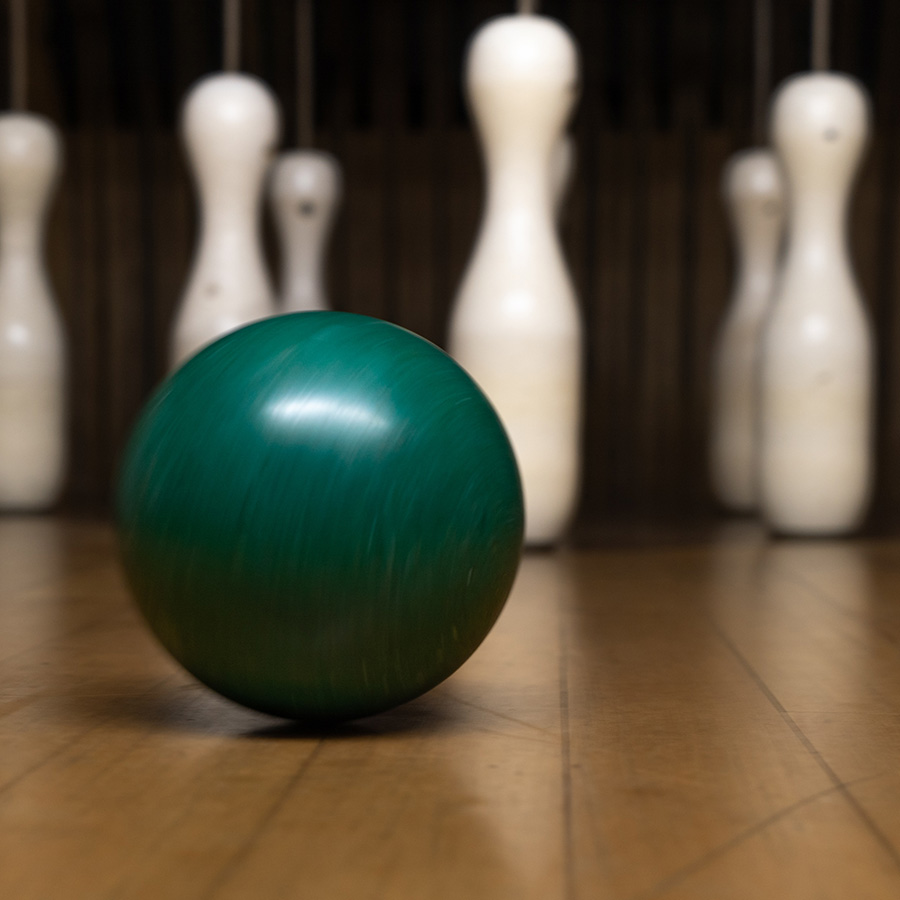 Kugel mit Kegeln im Hintergrund Eine grüne Bowlingkugel rollt auf einer Holzbahn zu weißen Kegel, die im Hintergrund stehen.