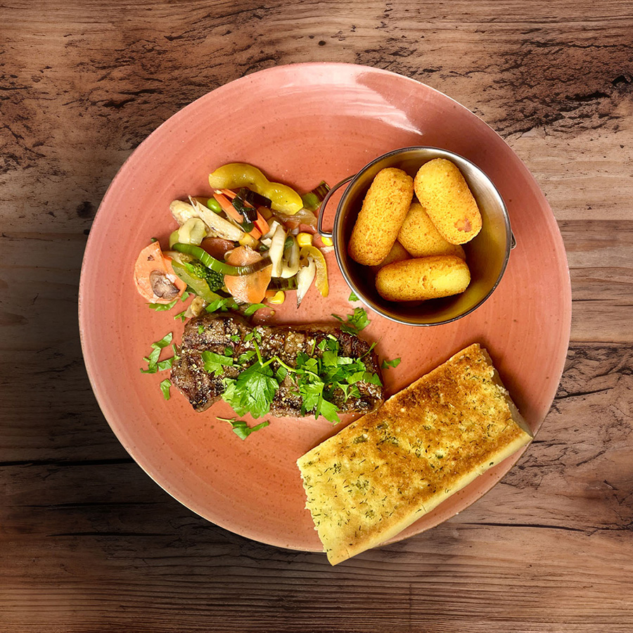 Close up of a brown hardwood table with a blurred background Ein Teller mit gegrilltem Fleisch, Gemüse, Knoblauchbrot und knusprigen Kroketten auf einem rustikalen Tisch.