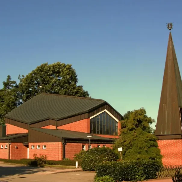 Restaurants_Hambühren_hero Moderne Kirche mit markantem spitzen Turm und roter Ziegelfassade, umgeben von Bäumen und Grünflächen.
