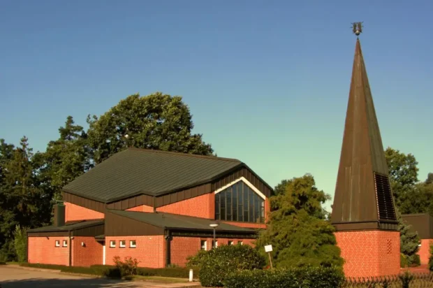 Moderne Kirche mit markantem spitzen Turm und roter Ziegelfassade, umgeben von Bäumen und Grünflächen.