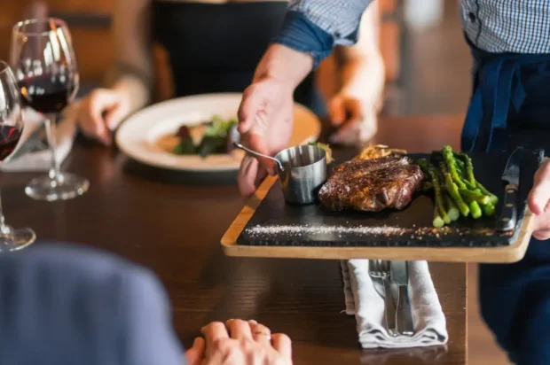 Hermannsburg_text Eine Bedienung serviert ein gebratenes Steak mit grünem Spargel und Soße auf einem schwarzen Schieferbrett im Restaurant.